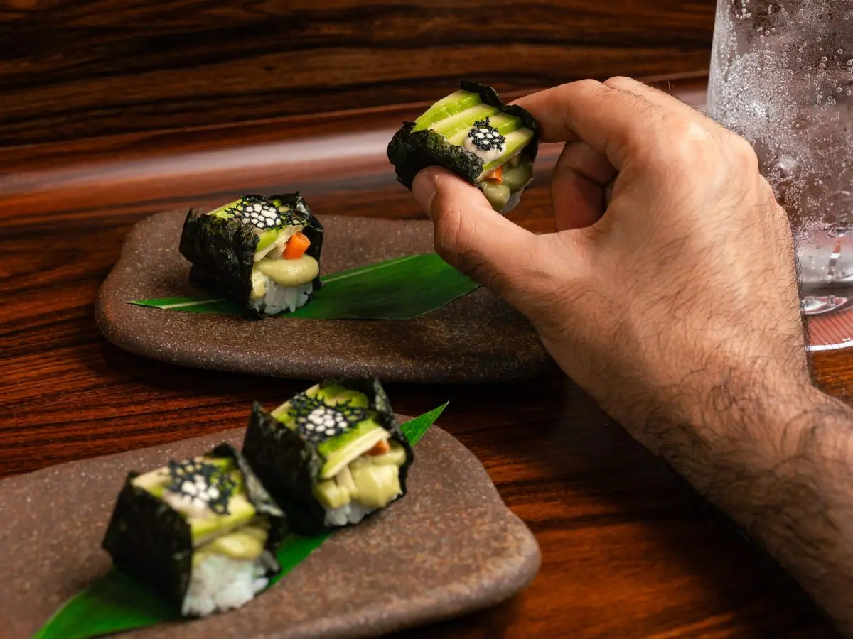 A hand lifting a maki from a stone plate at the counter